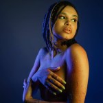 bare-chested african american woman with dreadlocks standing in colorful neon body paint, covering breast with hands and looking away on blue background with yellow lighting effect