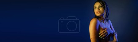 Téléchargez les photos : Charmante et nue femme afro-américaine avec dreadlocks, en peinture au néon coloré couvrant la poitrine avec les mains et regardant loin sur fond bleu avec effet d'éclairage jaune, bannière - en image libre de droit