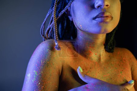 Teilaufnahme einer jungen und barbusigen afrikanisch-amerikanischen Frau in leuchtender und bunter Neon-Körperfarbe, die die Brust mit Händen auf blauem Hintergrund mit gelbem Lichteffekt bedeckt