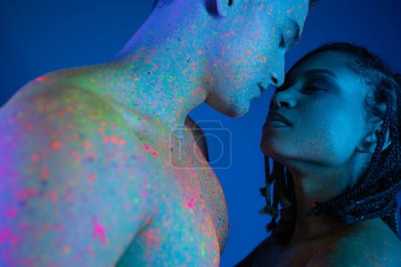 low angle view of young and sensual interracial couple with bare shoulders, in colorful neon body paint standing face to face with closed eyes on blue background with cyan lighting