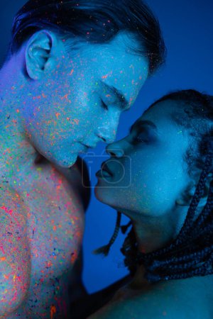 impassioned interracial couple standing face to face with closed eyes, bare-chested man and african american woman with dreadlocks on blue background with cyan lighting