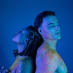 young interracial couple in colorful neon body paint standing back to back with closed eyes, nude african american woman covering breast near muscular man on blue background with cyan lighting