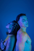 young interracial couple in colorful neon body paint standing back to back with closed eyes, nude african american woman covering breast near muscular man on blue background with cyan lighting t-shirt #658776646