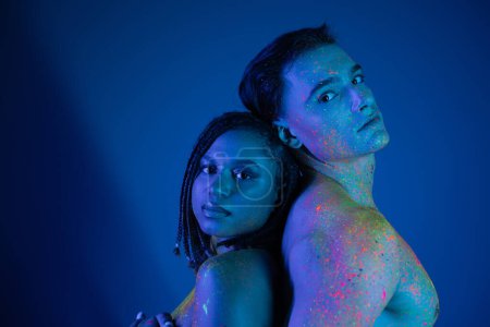 young multiethnic couple with naked shoulders, bare-chested, in colorful body paint looking at camera while posing on blue background with cyan lighting