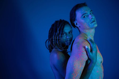 junges multikulturelles Paar in bunter Neon-Körperfarbe mit Blick in die Kamera auf blauem Hintergrund mit Cyanbeleuchtung, afrikanisch-amerikanische Frau umarmt hemdlosen Mann mit muskulösem Oberkörper
