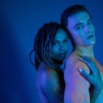 sexy interracial couple in colorful neon body paint looking at camera on blue background with cyan lighting, nude african american woman embracing shirtless man with muscular body