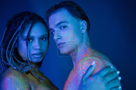 Photo for Youthful multicultural couple with bare shoulders, in colorful neon body paint looking at camera on blue background with cyan lighting, charismatic man and african american woman with dreadlocks - Royalty Free Image