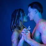 side view of interracial couple in colorful neon body paint standing with clenched hands, african american woman with dreadlocks and shirtless muscular man on blue background with cyan lighting