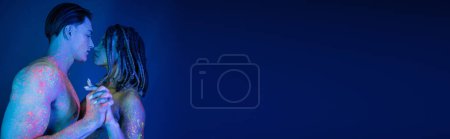 Téléchargez les photos : Homme musclé torse nu et femme afro-américaine avec dreadlocks regardant les uns les autres et debout avec les mains serrées dans de la peinture au néon colorée sur fond bleu avec éclairage cyan, bannière - en image libre de droit
