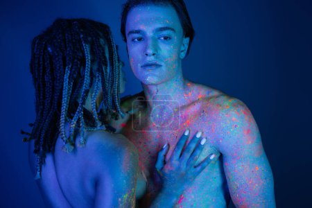 pareja interracial joven y desnuda en colorida pintura corporal de neón, mujer afroamericana con rastas cerca de hombre sin camisa con cuerpo muscular sobre fondo azul con iluminación cian