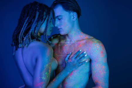 intimer Moment eines gemischtrassigen Paares in bunter Neon-Körperfarbe, nackte afrikanisch-amerikanische Frau, die die nackte Brust eines schulterfreien muskulösen Mannes auf blauem Hintergrund mit Cyan-Beleuchtung berührt