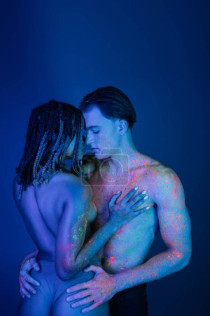 intimate moment of interracial couple in colorful neon body paint, confident shirtless man embracing sexy buttocks of young african american woman on blue background with cyan lighting