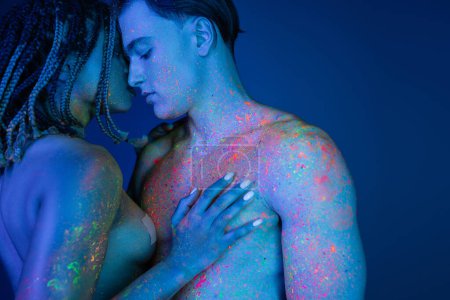 intimer Moment eines leidenschaftlichen multikulturellen Paares auf blauem Hintergrund mit cyanfarbener Beleuchtung, nackte afrikanisch-amerikanische Frau, die die Brust eines hemdlosen Mannes mit muskulösem Oberkörper berührt
