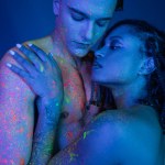 romantic multicultural couple in colorful body paint embracing on blue background with cyan lighting, nude african american woman with dreadlocks and shirtless man with muscular body