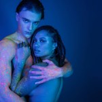 multicultural couple in colorful neon body paint embracing and looking at camera on blue background with cyan lighting, shirtless man with muscular body and african american woman with dreadlocks