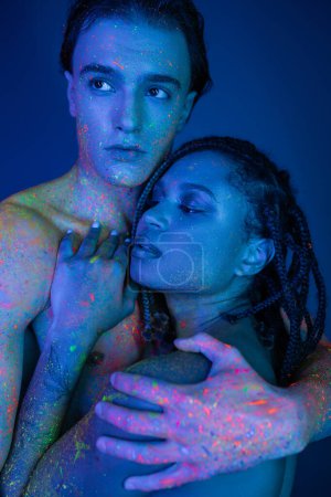 pareja interracial juvenil en colorida pintura corporal de neón abrazando sobre fondo azul con iluminación cian, hombre carismático sin camisa y mujer afroamericana con rastas