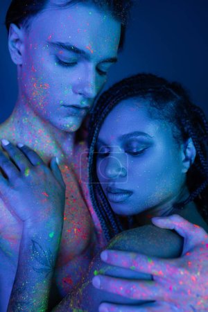 junges und sinnliches gemischtrassiges Paar in farbenfroher Körperfarbe, das sich auf blauem Hintergrund mit Cyanbeleuchtung umarmt, barbusiger Mann und afrikanisch-amerikanische Frau mit Dreadlocks und nackten Schultern