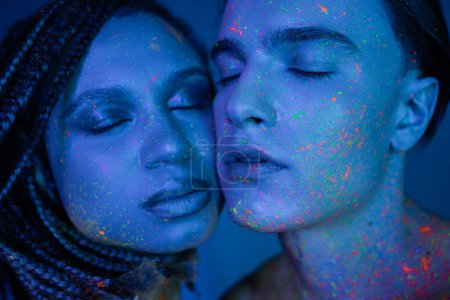 Porträt eines jungen multikulturellen Paares in bunter Neon-Körperfarbe, das mit geschlossenen Augen auf blauem Hintergrund mit Cyanbeleuchtung posiert, charismatischer Mann und faszinierende Afroamerikanerin