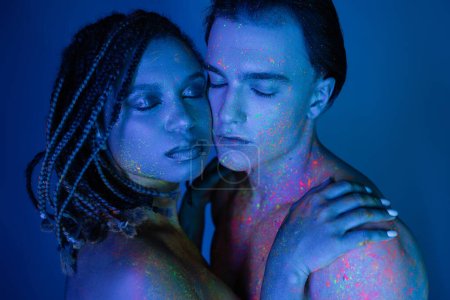 jeune couple interracial debout les yeux fermés sur fond bleu avec éclairage cyan, captivante femme afro-américaine embrassant les épaules nues de l'homme charismatique