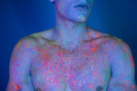 vista parcial del hombre sin camisa, con el pecho desnudo con el cuerpo muscular posando en multicolor y brillante pintura de cuerpo de neón, mientras que de pie sobre fondo azul con efecto de iluminación cian