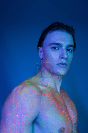 portrait of youthful and handsome shirtless man in multicolored neon body paint looking at camera while standing on blue background with cyan lighting effect