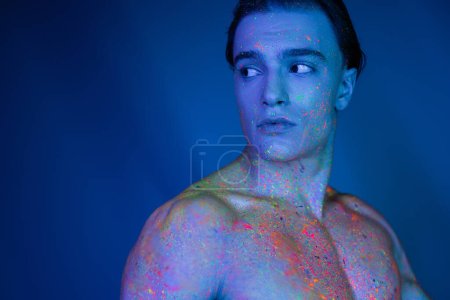 jugendlicher, auffälliger und hemdloser Mann mit muskulösem Körper in leuchtender und bunter Neon-Körperfarbe, der vor blauem Hintergrund mit Cyan-Lichteffekt wegschaut