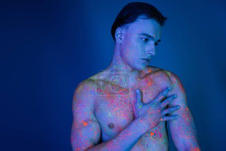 schöner junger, hemdloser Mann mit muskulösem Oberkörper, in bunter Neon-Körperfarbe, der im Stehen auf blauem Hintergrund mit Cyan-Lichteffekt die nackte Brust berührt