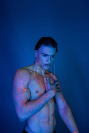 Jugendlicher hemdloser Mann mit muskulösem Oberkörper, der die nackte Brust berührt, während er in leuchtend bunter Neon-Körperfarbe auf blauem Hintergrund mit Cyan-Lichteffekt steht