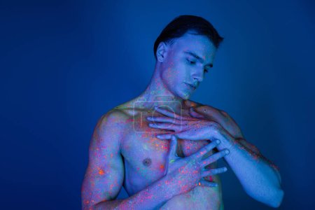 junger hemdloser Mann mit muskulösem Körper, der die nackte Brust berührt, während er in leuchtender und bunter Neon-Körperfarbe auf blauem Hintergrund mit Cyan-Lichteffekt posiert