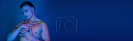 jugendlicher und charismatischer hemdloser Mann in heller Neon-Körperfarbe, der muskulösen Körper berührt, während er auf blauem Hintergrund mit Cyan-Lichteffekt posiert, Banner