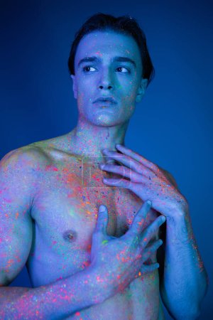 hombre joven, sin camisa y llamativo posando en vibrante pintura de cuerpo de neón colorido, tocando el pecho desnudo y mirando hacia otro lado sobre fondo azul con efecto de iluminación cian