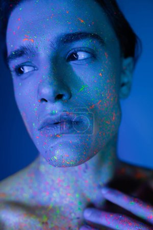 Nahaufnahme Porträt eines jugendlichen und gut aussehenden Mannes in leuchtender und mehrfarbiger Neon-Körperfarbe, der auf blauem Hintergrund mit Cyan-Lichteffekt wegschaut