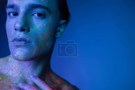 retrato de un joven guapo con expresión facial confiada posando en pintura corporal de neón multicolor mientras mira la cámara sobre fondo azul con efecto de iluminación cian