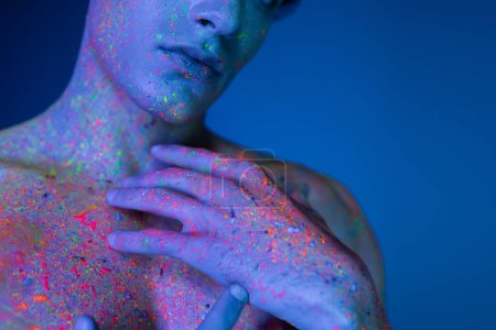 Photo for Cropped view of shirtless man holding hands near bare chest while posing in colorful and vibrant neon body paint on blurred and blue background with cyan lighting effect - Royalty Free Image
