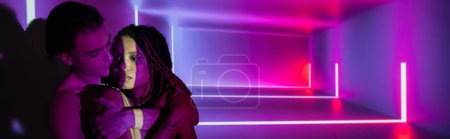 youthful and impassioned multicultural couple, sexy african american woman and youthful man embracing while standing on abstract purple background with neon rays and lighting effects, banner