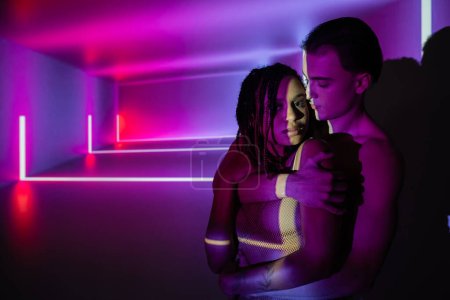 young and confident man embracing captivating african american woman with dreadlocks on abstract purple background with neon rays and lighting effects