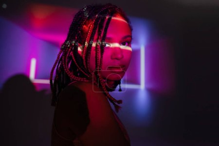 portrait of magnetic and appealing african american woman with dreadlocks looking at camera on abstract black and purple background with neon rays and lighting effects