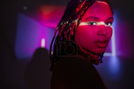 retrato de mujer afroamericana atractiva y joven con rastas elegantes mirando a la cámara mientras está de pie sobre fondo púrpura abstracto con rayos de neón y efectos de iluminación