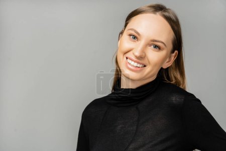 Porträt einer fröhlichen blonden Frau mit natürlichem Make-up, die ein stylisches schwarzes Kleid trägt und in die Kamera schaut, während sie isoliert auf grau mit Kopierraum steht 
