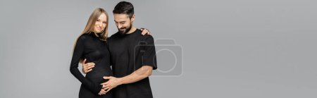 Cheerful bearded man in black t-shirt touching belly of fair haired and pregnant stylish wife in dress and hugging isolated on grey, growing new life concept, copy space, banner 