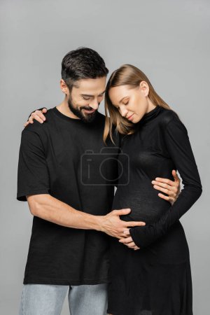 Smiling man in black t-shirt and jeans touching belly of pregnant and stylish wife while standing and hugging each other isolated on grey, growing new life concept