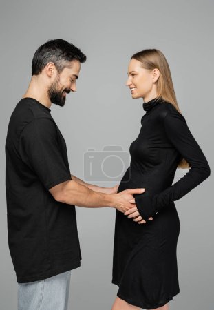Téléchargez les photos : Vue latérale de l'homme barbu positif en t-shirt noir touchant le ventre de la femme enceinte et blonde élégante en robe et debout ensemble isolé sur gris, croissant nouveau concept de vie - en image libre de droit