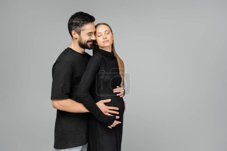 Joyful bearded man in t-shirt hugging belly of stylish and relaxed pregnant wife in black dress while standing with closed eyes isolated on grey, growing new life concept
