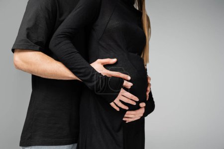 Ausgeschnittene Ansicht eines fürsorglichen Mannes im schwarzen T-Shirt, der den Bauch einer stilvollen schwangeren Frau im Kleid umarmt und isoliert auf grauem, wachsendem neuen Lebenskonzept steht