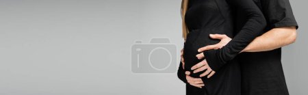 Ausgeschnittene Ansicht eines Mannes im T-Shirt, der den Bauch einer hochschwangeren Frau in schwarzem Kleid umarmt und berührt und isoliert auf grauem, wachsendem neuen Lebenskonzept steht, Banner 