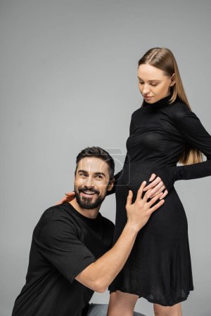 Smiling bearded man in t-shirt listening belly of trendy and pregnant wife with natural makeup in stylish black dress while standing isolated on grey, growing new life concept