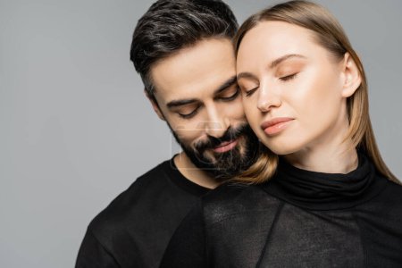 Portrait of relaxed and bearded man in black t-shirt standing next to wife with fair haired wife with natural makeup and closed eyes isolated on grey, husband and wife relationship concept