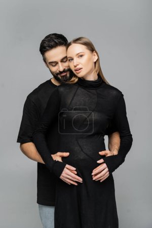 Bearded man in black t-shirt touching belly of stylish and pregnant fair haired wife in dress and standing together isolated on grey, concept of pregnancy and birth of child