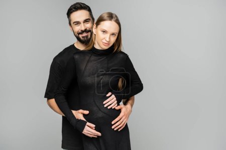 Hombre positivo y barbudo en camiseta negra abrazando a la esposa de moda y embarazada en vestido negro y mirando a la cámara mientras está de pie aislado en gris, concepto de nacimiento del niño