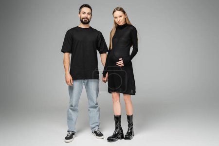 Full length of stylish and fair haired pregnant woman in black dress holding hand of bearded husband and looking at camera on grey background, concept of expecting parents